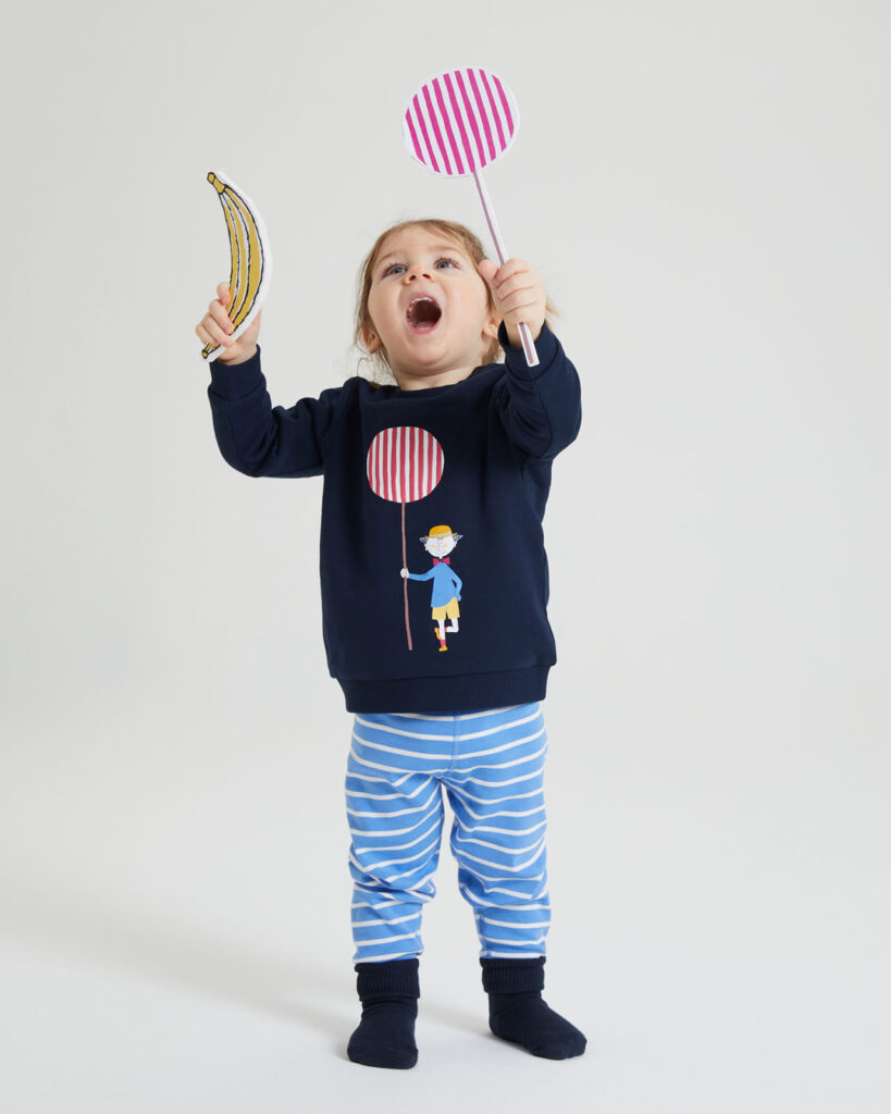 Child wearing sweater with krakel spektakel illustration, striped pants and holding a lollipop and banana